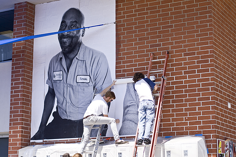 Senior, Evan Cassell and Contemporary photo T.A. Seth Johnson add another portrait to the SUB wall while other photo students paste portraits on the library. | Katie Juranek/The Chimes
