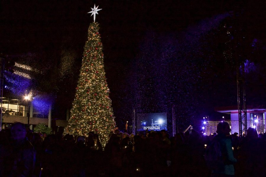 Biola students, faculty, parents, and alumni gather around the lit Christmas tree enjoying the light, fake snow, and fellowship